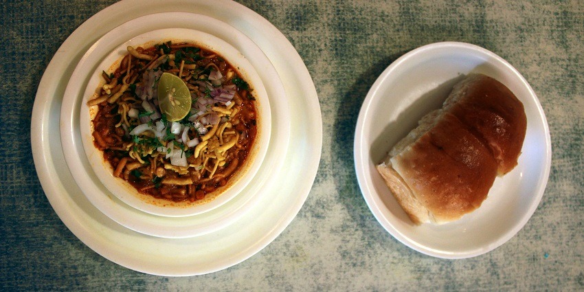 A bowl of spicy misal topped with farsan served with pav on the side.