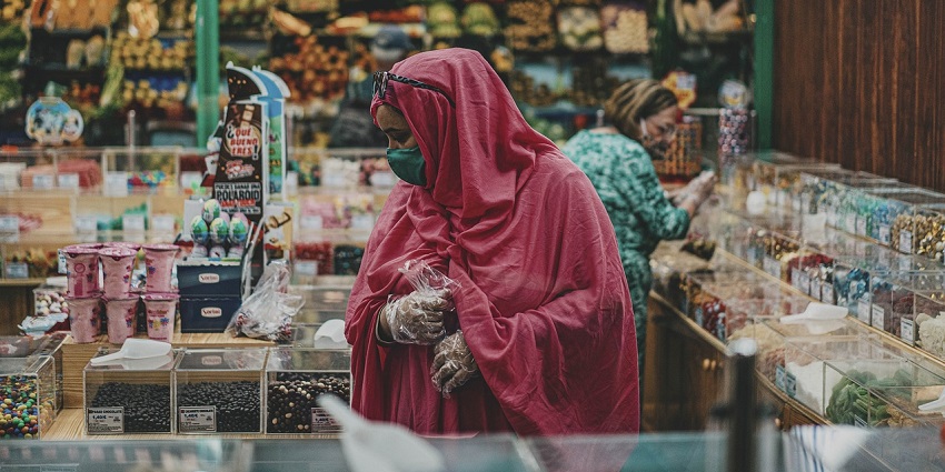 A woman wearing a face mask and hijab smiles while shopping in a vibrant market setting.