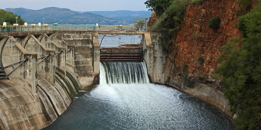 A dam with a large open sluice gate, releasing water into a river, surrounded by green hills