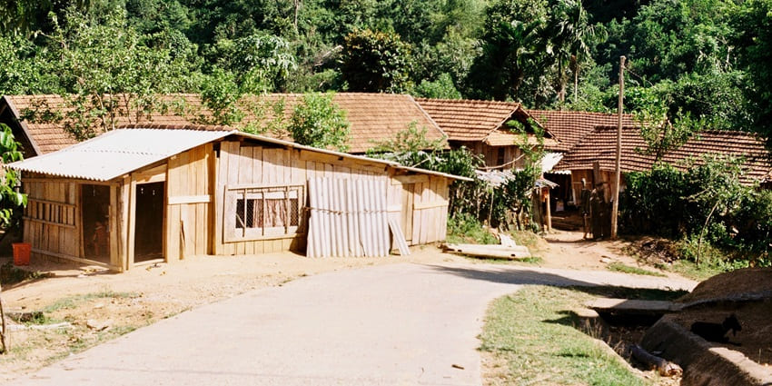 Rustic village houses with tiled and tin roofs surrounded by dense green forested hills.