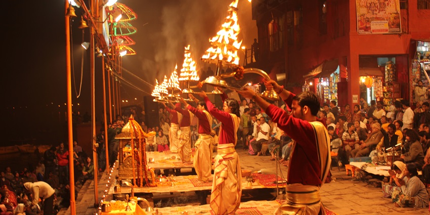 A picture of priests offering evening prayers at a river ghat in Madhya Pradesh, with an illuminated aarti thali