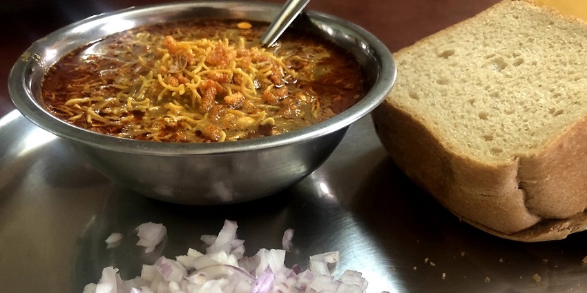 Spicy Kolhapuri misal served with two slices of bread, chopped onions and lime wedge