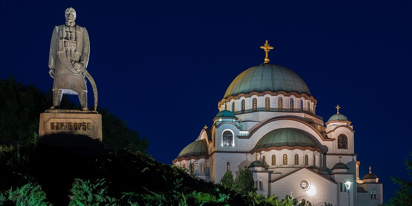 A nighttime picture of a Church in Serbia with a tall statue next to it, surrounded by greenery