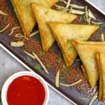 Five triangular-shaped snacks served on a black tray accompanied by a red dip