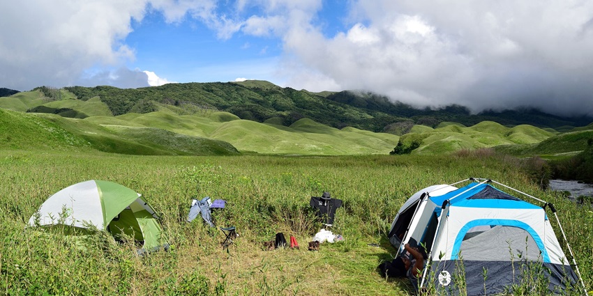 Multi-coloured tents set up for camping in a huge field with greenery and clouds.