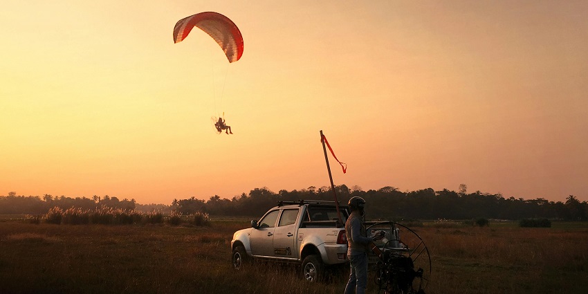 A person paragliding in the sunset with another man on the land standing near a car