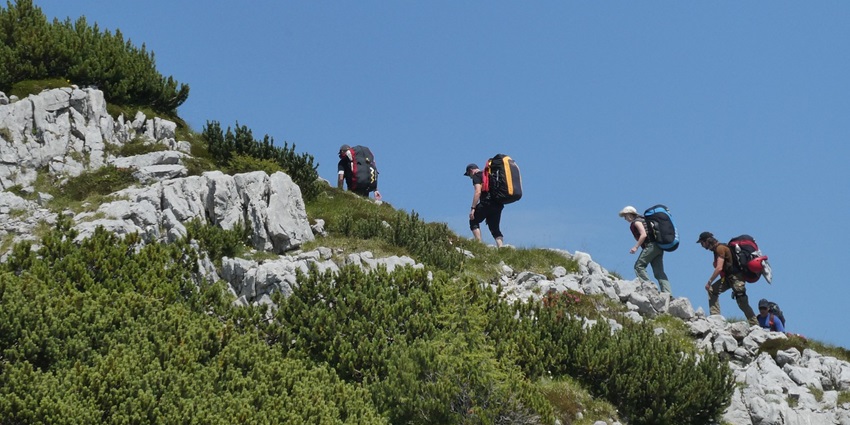 Tourists climbing a green and rocky mountain trail, carrying large backpacks with them.