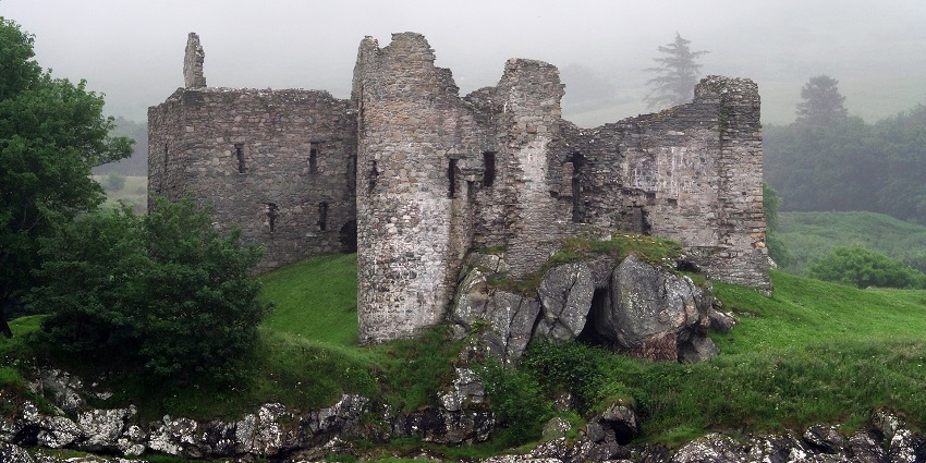 Wide view of an ancient stone castle ruin on a grassy hill under a cloudy sky