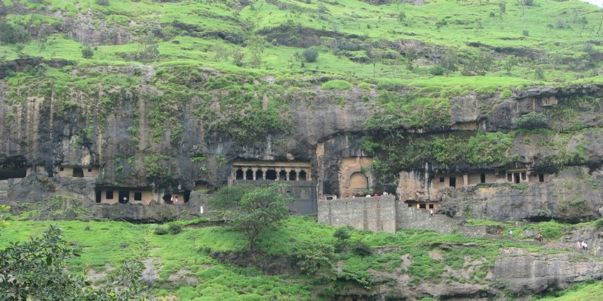 Ancient Buddhist caves carved into a hillside, with greenery and a mountain backdrop.