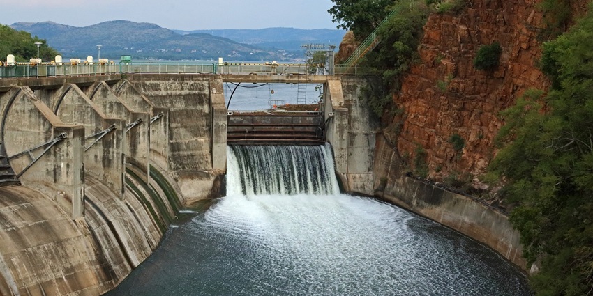 A large dam with open gates, letting water flow into a wide river, with green hills in the background