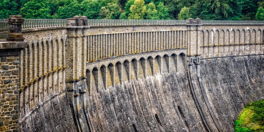 Concrete dam wall and barrier reflecting under a clear blue sky and azure waters.