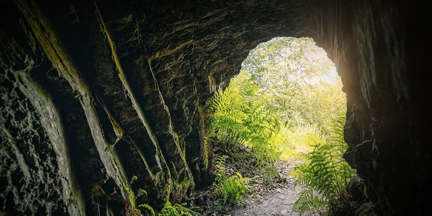 A dimly lit cave tunnel with stone walls leads to a bright light at the end, creating a sense of mystery.
