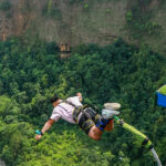 A man in mid-air during his bungee jump, with green hills and another man on the pavement