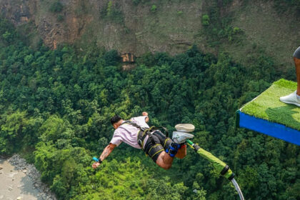 A man in mid-air during his bungee jump, with green hills and another man on the pavement