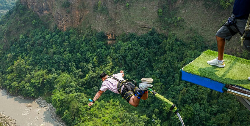 A man in mid-air during his bungee jump, with green hills and another man on the pavement