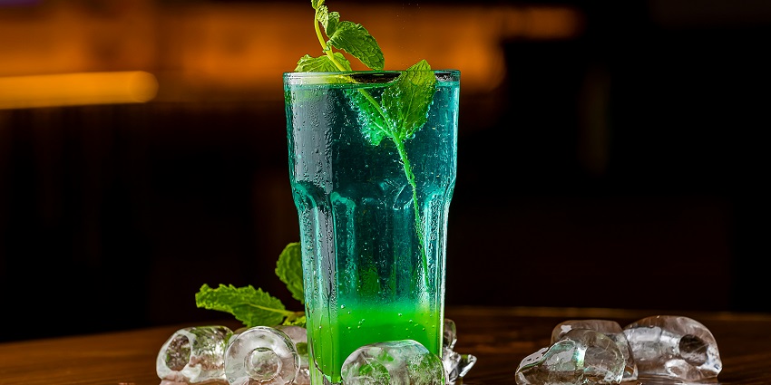 A lip-smacking view of a glass with green and blue drink served with ice on the side.