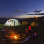 Bonfire and camping tents set up during the night with hills in the background.