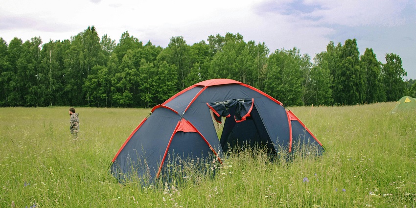 A blue tent is placed among the woods, and nearby it, there is a wooden picnic table.