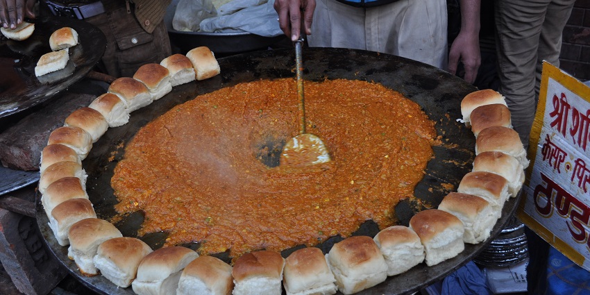 Vendor is cooking Bhaji on black round iron tawa surrounded by neatly arranged pavs.
