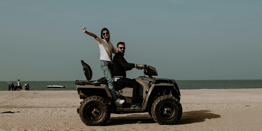 A couple is posing on a fancy motorbike on a beachside, with the lady standing on the bike.