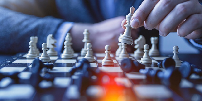 The image depicts a person moving a chess piece on a wooden board in an indoor setting
