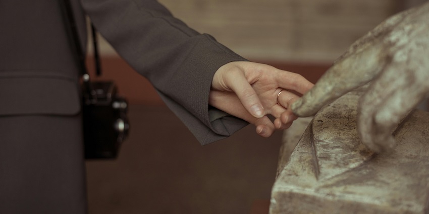 A woman's hand gently touching the fingers of an old, detailed and textured sculpture