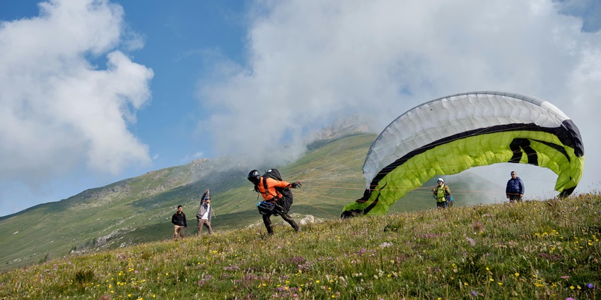 Paraglider preparing to take off on a grassy hilltop with puffy clouds and people staring.