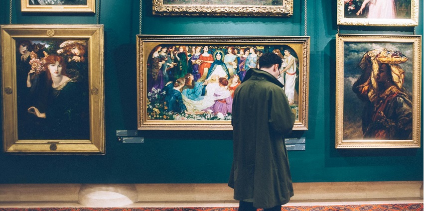 A man stands in front of a gallery wall, looking at a collection of framed paintings.
