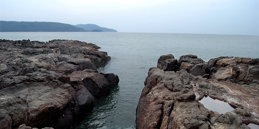 A rocky shoreline with dark, jagged rocks framing a narrow channel of ocean water.