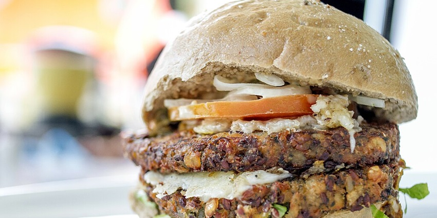 A double vegetable burger with a layer of tomato slices, onions, and lettuce is served.