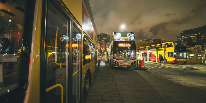 Two buses traveling on a road surrounded by greenery under a clear blue sky.