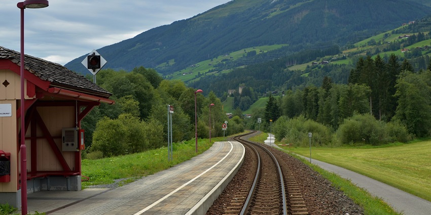 Train pulling into station with passengers disembarking, set against the scenic backdrop.