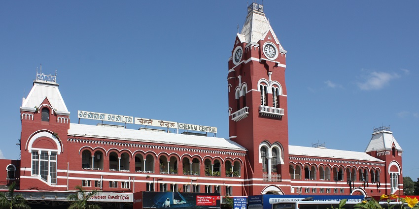 A busy Chennai train station with passengers, trains, and vendors under a clear blue sky.