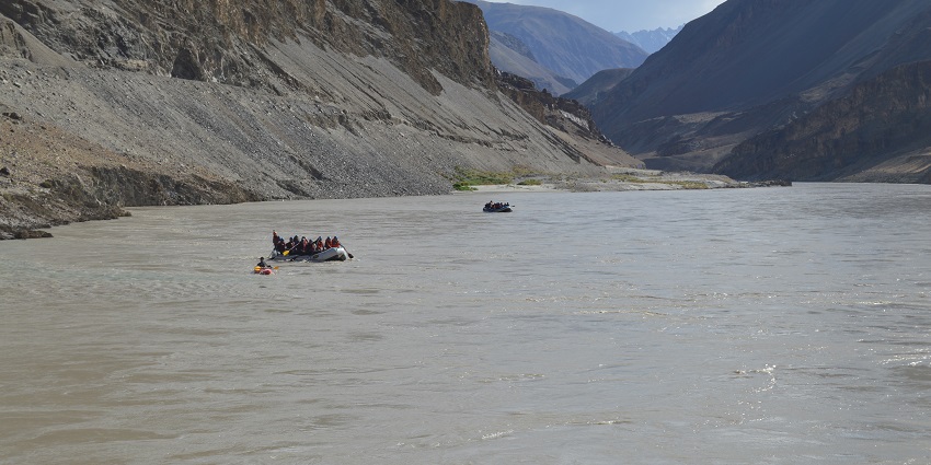 A picture of a river in Ladakh with multiple mountains visible in the background and people rafting in the river
