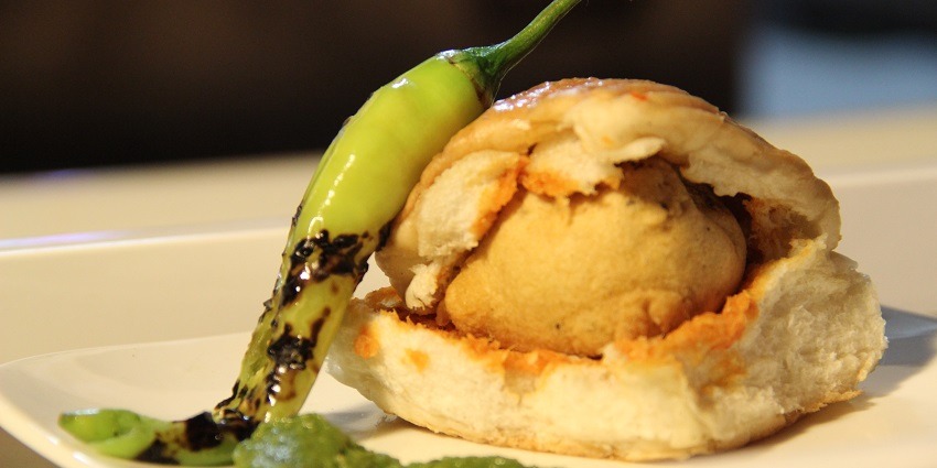 A person holding a plate of pav with a big vada inside and served with green chillies
