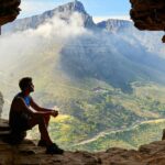 The image shows a man sitting inside a large cave in Maharashtra looking out.