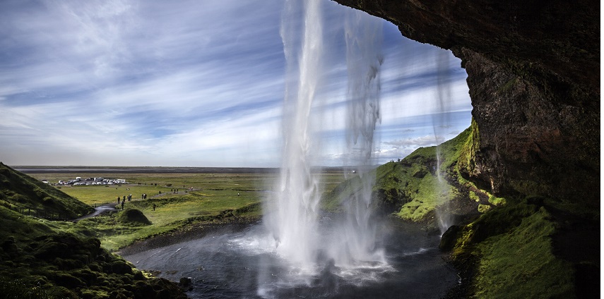 A waterfall flows over rocky ledges, surrounded by vibrant green foliage and sunlight.