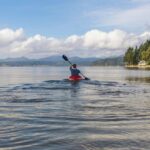 Solo kayaker paddling across a tranquil lake surrounded by green trees and distant hills.