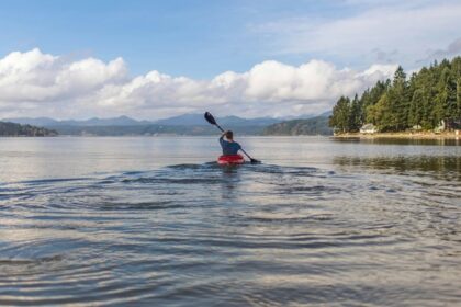 Solo kayaker paddling across a tranquil lake surrounded by green trees and distant hills.