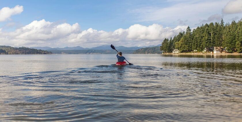 Solo kayaker paddling across a tranquil lake surrounded by green trees and distant hills.