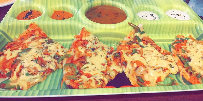 Pizza dosa topped with veggies and cheese served on a banana leaf with different chutneys