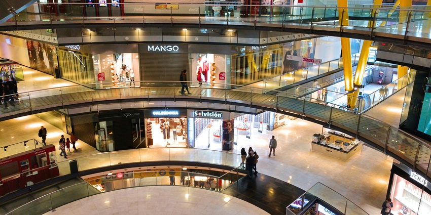 Multi-level shopping mall with glass railings, various brand stores, and few shoppers.