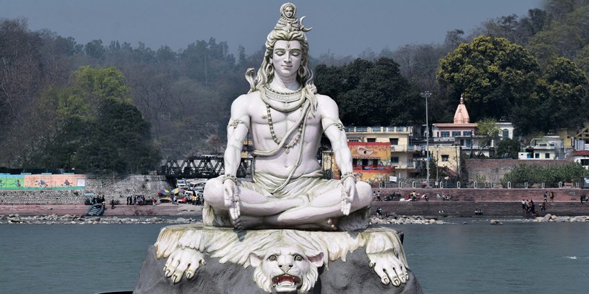 A statue of Lord Shiva meditating sitting on a huge rock with a serene river behind it
