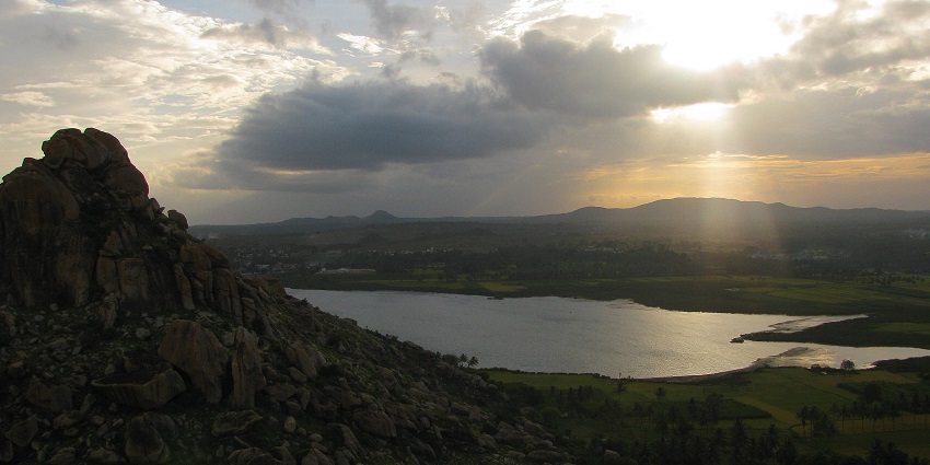 A rocky hilltop overlooks a lake and valley at sunset, with dramatic clouds and sunlight.