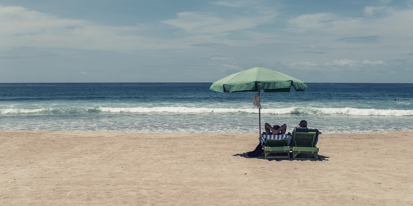 Two people sitting under an umbrella at Kuta Beach with gentle waves against clear sky, one of the popular places to visit in Kuta.