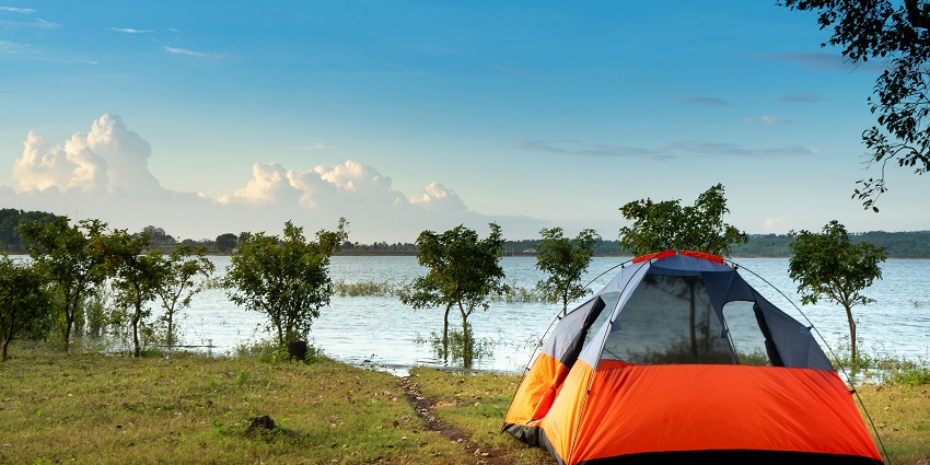 A green-coloured tent is placed on the ground near the bank of a lake while the sun is rising