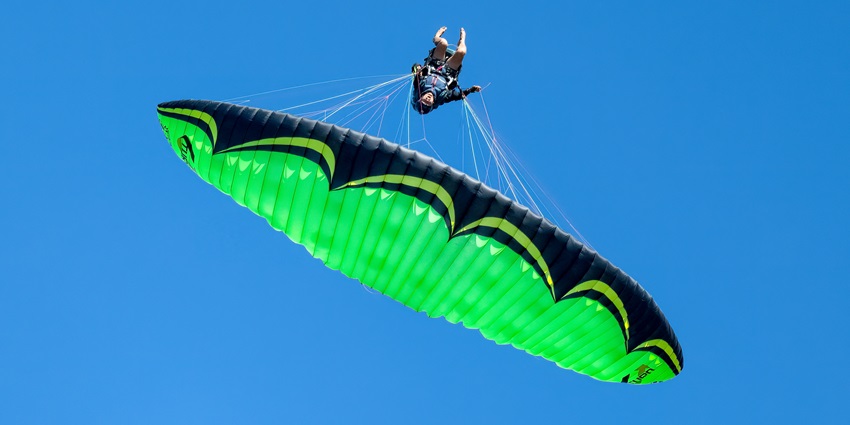 A paraglider performing an acrobatic infinity tumbling manoeuvre against a blue sky.