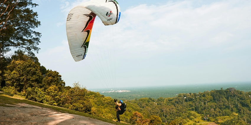Man gripping a parachute harness, preparing to takeoff or landing in the hilly area.