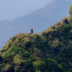 A tall, rocky pinnacle rises above lush green hills under a clear blue sky in Maharashtra.