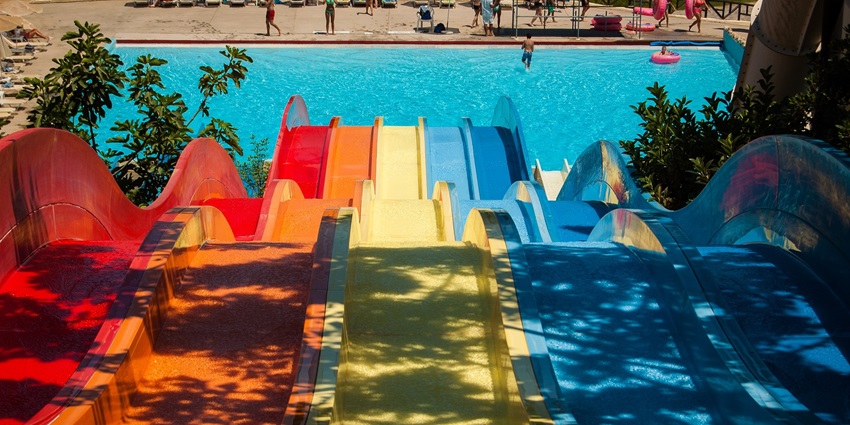 A colourful water slide in a sunlit water park, surrounded by greenery and clear blue sky.